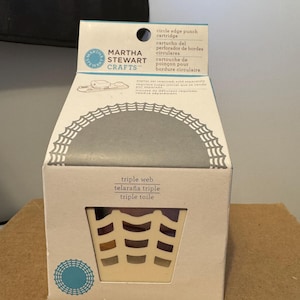Op de afbeelding: Een Martha Stewart Crafts cirkelvormige pons cartridge doos. De witte doos heeft een decoratief webdesign en de woorden "triple web" in het Engels, Spaans en Frans. De doos bevat een pons cartridge.