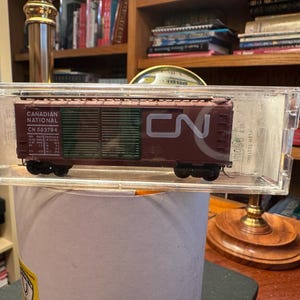 Pode incluir: Um modelo de vagão de trem numa caixa de exibição transparente. O vagão é marrom com detalhes verdes e o texto "CANADIAN NATIONAL" e "CN 583794". Possui rodas pretas e está exposto numa mesa.