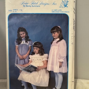 Puede incluir: Un patrón de costura para un vestido de cuello smock para niñas. El patrón se llama "Amy-Lori" y es de Briar Patch Designs, Inc. El patrón incluye tallas de 6 meses a 4 años y de 5 a 8 años. La imagen muestra a tres niñas usando el vestido en diferentes colores.