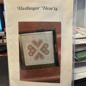 Peut inclure: Un motif de broderie blanc et brun pour un modèle de Hardanger Hearts. Le motif est encadré dans un cadre en bois brun et montre un trèfle à quatre feuilles avec des cœurs au centre. Le motif est de Threads and Things, Karen R. Buell, 303 Lakeview Dr., Hoyt Lakes, MN 55750.