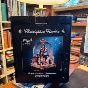 May include: A black box featuring a Christopher Radko Nutcracker Suite snowglobe. The box has text that reads "Musical! Rotates with blower!" and "LIMITED EDITION SERIES PLAYS, Nutcracker Suite". The snowglobe is visible on the box.