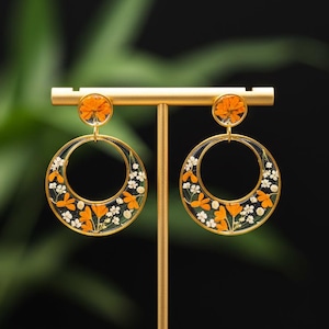 Puede incluir: Pendientes de aro dorados con detalles florales naranjas y blancos. Los pendientes presentan un diseño circular con un círculo más pequeño lleno de flores en la parte superior. Las flores están engarzadas en resina transparente.