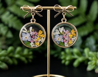 Op de afbeelding: Goudkleurige oorbellen met een strikdesign. Elke oorbel heeft een heldere, cirkelvormige hanger gevuld met kleurrijke gedroogde bloemen. De oorbellen worden gepresenteerd op een goudkleurige standaard tegen een achtergrond van groen gebladerte.