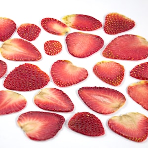 Puede incluir: Vista aérea de fresas en rodajas sobre un fondo blanco. Las fresas son de un rojo vibrante con semillas visibles y un interior claro. Algunas rodajas están enteras, otras cortadas por la mitad, mostrando la textura de la fruta.