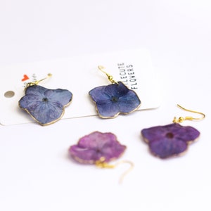 May include: Earrings featuring pressed blue and purple hydrangea flowers with gold-coloured edges and ear wires. The earrings are displayed on a white card with the text "LITTLE CUTE FLOWERS".