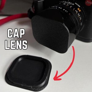 Puede incluir: Una tapa de lente de cámara negra con las palabras "CAP LENS" impresas en blanco en el lateral. La tapa de la lente está sobre una superficie blanca. Se puede ver una lente de cámara negra con un anillo plateado en el fondo.