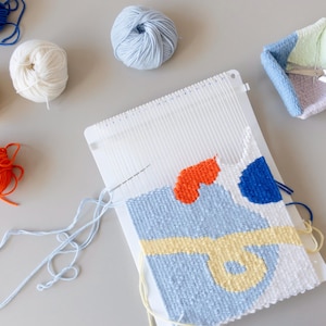 Peut inclure: Un métier à tisser blanc avec un motif tissé bleu, jaune et orange. Il y a des pelotes de laine de différentes couleurs, notamment bleu, blanc, jaune et orange, et une paire de ciseaux dans un petit panier tissé.
