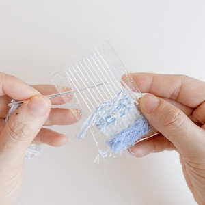 Puede incluir: Primer plano de un proyecto de tejido en curso. Un telar de plástico transparente sostiene hilos blancos, con una sección parcialmente tejida en tonos azules y blancos. Una aguja se inserta a través de los hilos.