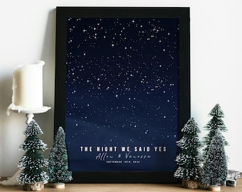 Puede incluir: Una impresión enmarcada con un cielo nocturno azul oscuro lleno de estrellas blancas. Se muestra el texto "THE NIGHT WE SAID YES", junto con nombres y una fecha. En primer plano, hay árboles de Navidad en miniatura y una vela.