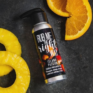 Our edible massage oil "Island Passion" is shown propped with pineapple rings and orange slices on a dark background. Flavored with mango, pineapple, and orange, this tropical massage oil is a delicious and natural addition to intimacy.