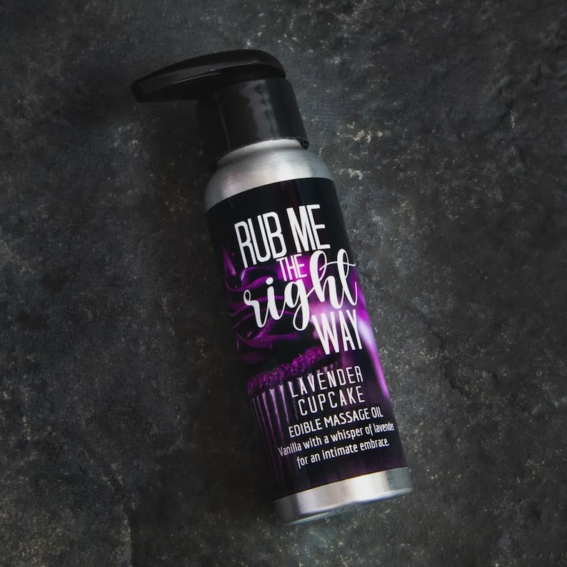 May include: A silver bottle with a black pump top, featuring the text "RUB ME the right WAY" in white and purple. The label reads "LAVENDER CUPCAKE EDIBLE MASSAGE OIL" with additional text. The bottle is on a dark gray surface.