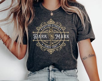 May include: Dark gray t-shirt featuring a gold and white graphic design for a tattoo studio. The design includes the text "Tattoo Studio Dark Mark Since 1967" in a circular and ornate layout. The shirt is a crew neck.