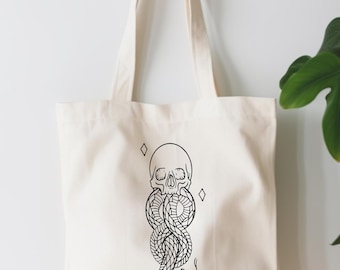 May include: Cream-colored tote bag featuring a black illustration of a skull intertwined with a snake. Two diamond shapes flank the skull. The bag has long handles for carrying over the shoulder.