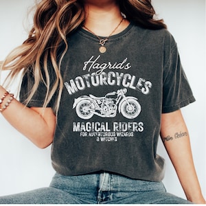 Pode incluir: Uma camiseta cinza escura com um design gráfico branco que diz "Hagrid's Motorcycles" com uma ilustração de motocicleta vintage e o texto "Magical Riders for Adventurous Wizards & Witches".