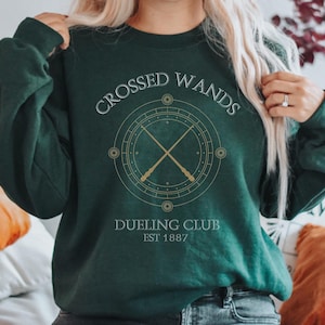 May include: A dark green sweatshirt with the text "CROSSED WANDS DUELING CLUB EST 1887" and a graphic of two crossed wands inside a circle.