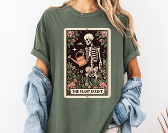 May include: A green t-shirt with a graphic of a skeleton watering plants. The graphic is in a tarot card style with the text "The Plant Parent".