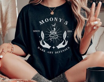 May include: Black t-shirt with a white graphic design that says "Moony's Since 1993 Dark Art Defenses". The graphic features a snake wrapped around a crescent moon with stars and leaves.