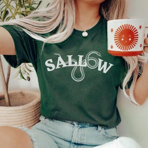 May include: A dark green t-shirt with a white snake and the word "SALL0W" printed on the front. The person wearing the shirt is holding a red mug with a white sun design.
