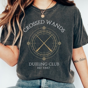 Può includere: Una maglietta grigia scura con il testo "Crossed Wands Dueling Club Est 1887" e una grafica di due bacchette incrociate all'interno di un cerchio con stelle e altri simboli.