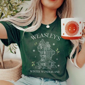 May include: Dark green t-shirt with the text "WEASLEY'S WINTER WONDERLAND" and a detailed line drawing of a whimsical house. A white and orange mug with a sun design is held in the hand.