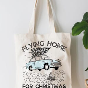 以下が含まれることがあります： 「FLYING HOME FOR CHRISTMAS」の文字が入ったクリーム色のトートバッグ。テキストの下には、屋根にクリスマスツリーを載せた水色の車が描かれています。バッグにはクリーム色のハンドルが2つ付いています。