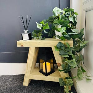 May include: A small, light-coloured wooden side table with a black lantern and a faux plant. The lantern has a yellow candle inside. A black diffuser bottle with reeds sits on the table. The table is 61 cm tall.
