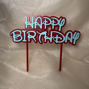 May include: A red cake topper with the words "HAPPY BIRTHDAY" in a stylized font. The text is in a light blue colour. The topper has two red sticks for insertion into a cake.