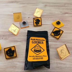 May include: A collection of acrylic mystery pins with various designs, including cat and fox faces, and a black bag with the text "Studio Wednesday GOOBER." The pins are in gold, black, and yellow.