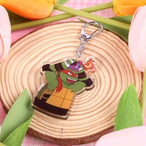 Puede incluir: Un llavero de acrílico transparente con un personaje de dibujos animados verde, naranja y morado con un pañuelo rojo y un pequeño personaje morado en la cabeza. El llavero está hecho de acrílico transparente y tiene un anillo de metal plateado.