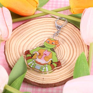 May include: A green and orange Teenage Mutant Ninja Turtle character holding a pink cat keychain. The keychain is attached to a silver key ring.