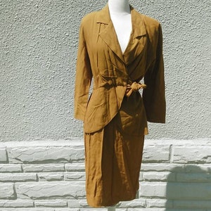 May include: A mustard yellow dress suit with a long-sleeved jacket and a skirt. The jacket has a V-neckline and a bow tie at the waist. The skirt is knee-length.