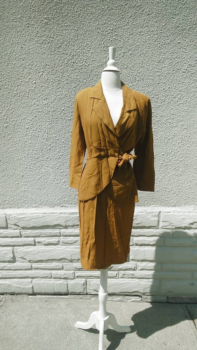 May include: A mustard yellow dress suit with a long-sleeved jacket and a skirt. The jacket has a V-neckline and a bow tie at the waist. The skirt is knee-length.