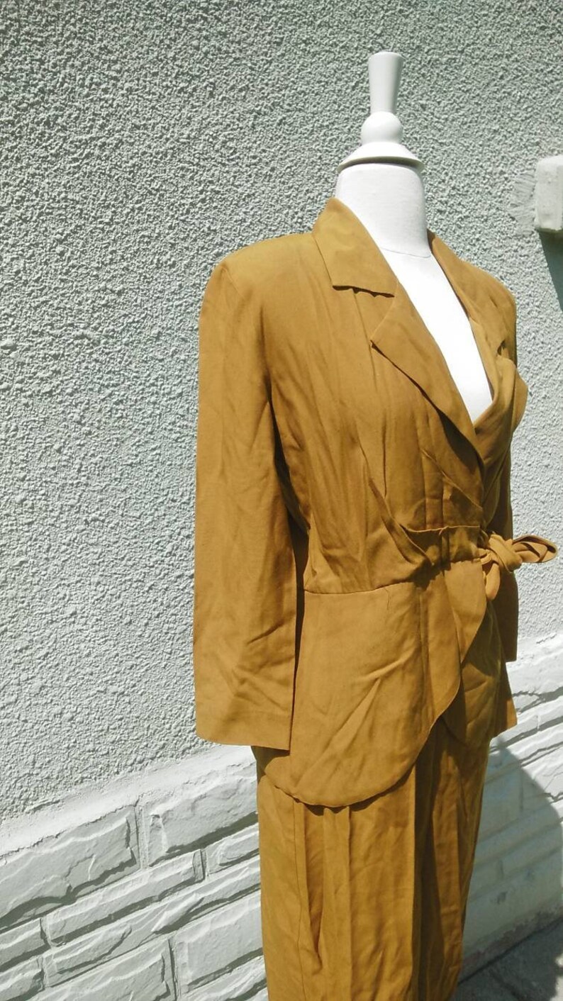 May include: A mustard yellow suit with a long-sleeved jacket and a skirt. The jacket has a wrap-style closure and a bow at the waist.