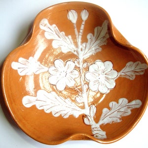 May include: A terracotta ceramic dish with a white floral design. The dish has a scalloped edge and is decorated with a stylized flower and leaf pattern.