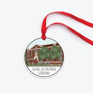 May include: A white round ornament with a red ribbon. The ornament features a cartoon illustration of a brick building with a sign that reads "US Army Recruiting and Retention Command Headquarters." The text "USARec Baton Rouge Louisiana" is printed below the illustration.