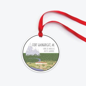 May include: A white round ornament with a red ribbon. The ornament features a cartoon illustration of a mountain range, a river, and a building with the text "FORT WAINWRIGHT, AK HOME OF AMERICA'S ARCTIC WARRIORS".
