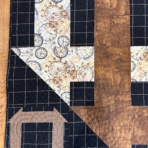 May include: A quilted textile with a patchwork design. The quilt features a black plaid pattern, sections with clock faces, and a brown, wood-grain-like pattern. The quilt has a geometric arrow and square design.