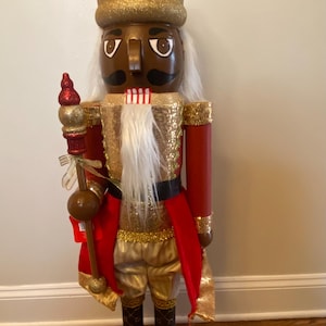 Puede incluir: Una figura de cascanueces de madera con una corona dorada, ropa roja y dorada y una barba blanca. El cascanueces sostiene un bastón dorado y rojo.