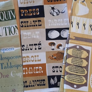 May include: Three rectangular kitchen towels featuring culinary themes. The left towel is olive green with text about measurement equivalents. The center towel is beige and black with cooking verbs and food illustrations. The right towel is yellow with kitchen tool graphics.
