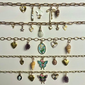 May include: Gold-toned charm necklaces with various pendants. Charms include hearts, stars, seashells, butterflies, and cameos. The necklaces are displayed on a white surface, showcasing the intricate details of each charm.