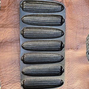 May include: A cast iron cornbread pan with six corn cob moulds. The pan is dark grey and has a handle with a hole for hanging. The moulds are detailed to resemble corn on the cob.