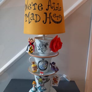 May include: A decorative lamp with a yellow lampshade, featuring the text "We're All Mad Here". The lamp is made of stacked teacups and saucers, decorated with Alice in Wonderland characters, a red rose, and a playing card.