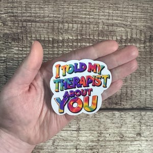 May include: A white sticker with the text "I TOLD MY THERAPIST ABOUT YOU" in colorful, rainbow-style lettering. The sticker is held in a person's hand against a wooden background.