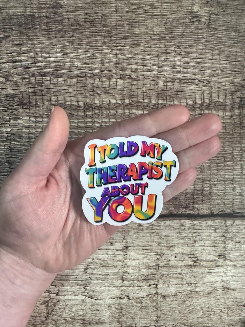 May include: A white sticker with the text "I TOLD MY THERAPIST ABOUT YOU" in colorful, rainbow-style lettering. The sticker is held in a person's hand against a wooden background.