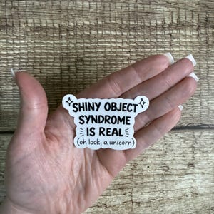 May include: A white sticker with black text that reads "SHINY OBJECT SYNDROME IS REAL (oh look, a unicorn)". The sticker has a starburst design on each side of the words "SHINY OBJECT". The sticker is held in a hand.