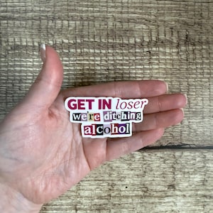 May include: A rectangular sticker with the text "GET IN loser we're ditching alcohol" in a mix of fonts and colors. The sticker is held in a hand against a wooden background.