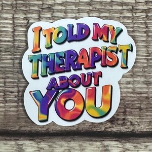 May include: A white sticker with the text "I TOLD MY THERAPIST ABOUT YOU" in colorful, tie-dye style lettering. The sticker is on a wooden surface.