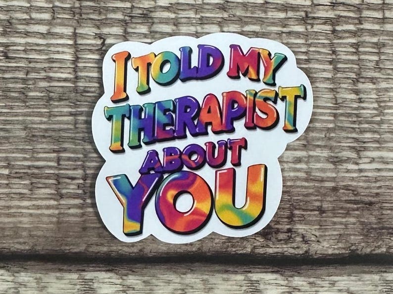 May include: A white sticker with the text "I TOLD MY THERAPIST ABOUT YOU" in colorful, tie-dye style lettering. The sticker is on a wooden surface.