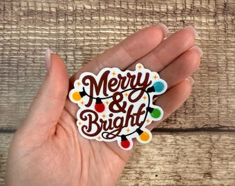 May include: A sticker with the words "Merry & Bright" in a decorative font, surrounded by a string of colorful Christmas lights. The sticker is held in a hand against a wooden background.