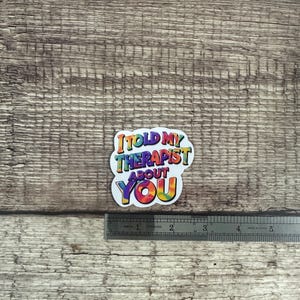 May include: A colorful sticker with the text "I told my therapist about you." The text is in a rainbow tie-dye design. A metal ruler is next to the sticker, showing inches.